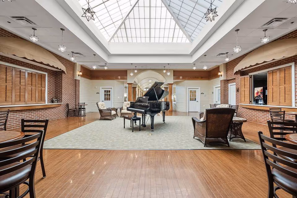 A spacious common area with a grand piano in the center on a large carpet. Surrounding the piano are wicker chairs and small tables. The room features wooden floors, brick walls with shuttered windows, and a large skylight ceiling with star-shaped pendant lights.