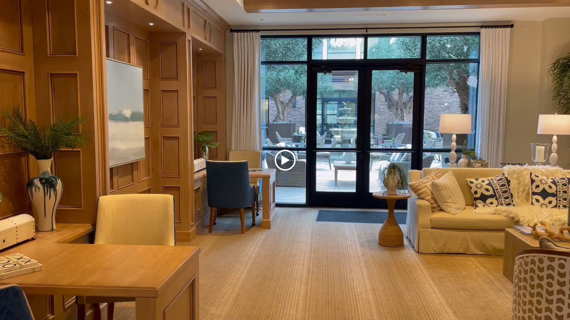 A cozy interior living area with wooden paneling on the left wall, a wooden table with chairs, a beige sofa adorned with patterned cushions and a fur throw, two table lamps, and a small round side table with a plant. Large glass doors at the back open to an outdoor patio with seating and greenery.