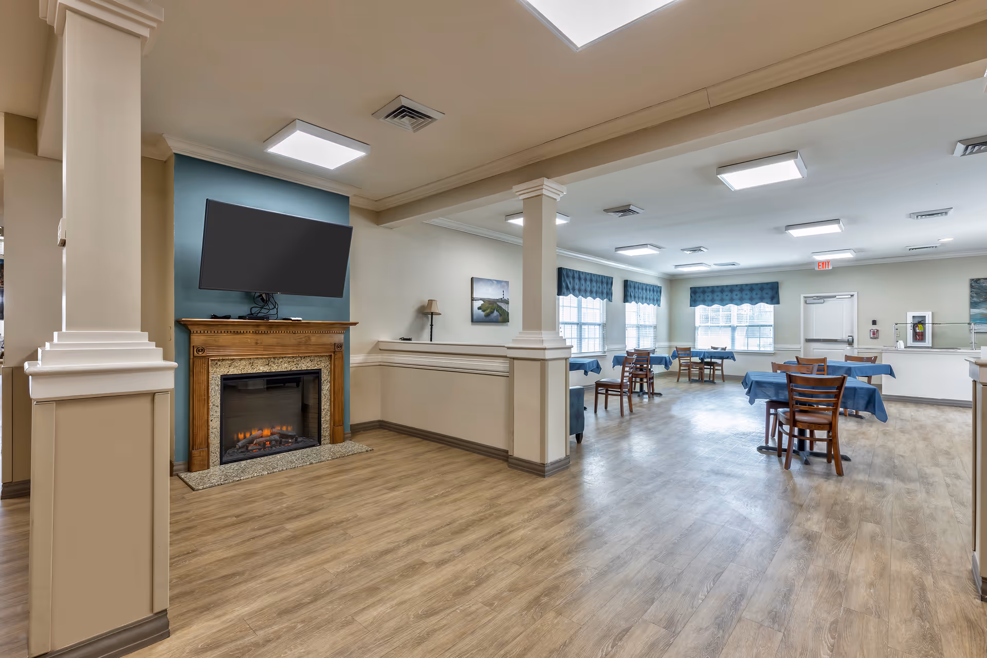 A spacious dining area with several wooden tables and chairs covered with blue tablecloths. The room features large windows with blue valances, a wooden floor, and a fireplace with a mounted flat-screen TV above it. The walls are painted in neutral tones with white trim and columns dividing the space.