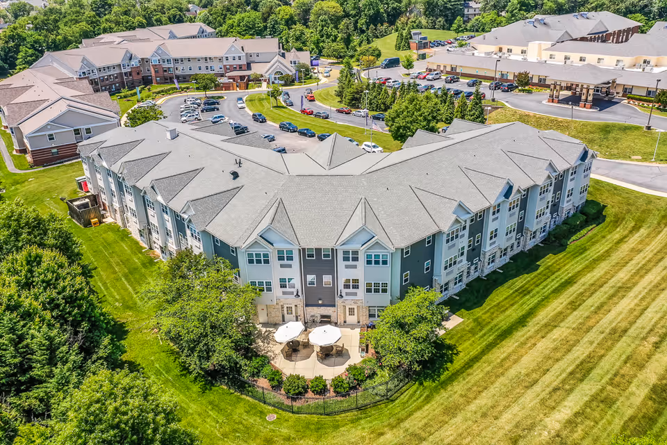 Aerial view of The Ridge at Sterling senior living facility showing multiple connected buildings with gray roofs, surrounded by well-maintained green lawns, trees, and parking areas with cars. There is a small fenced patio area with tables and umbrellas visible in the foreground.