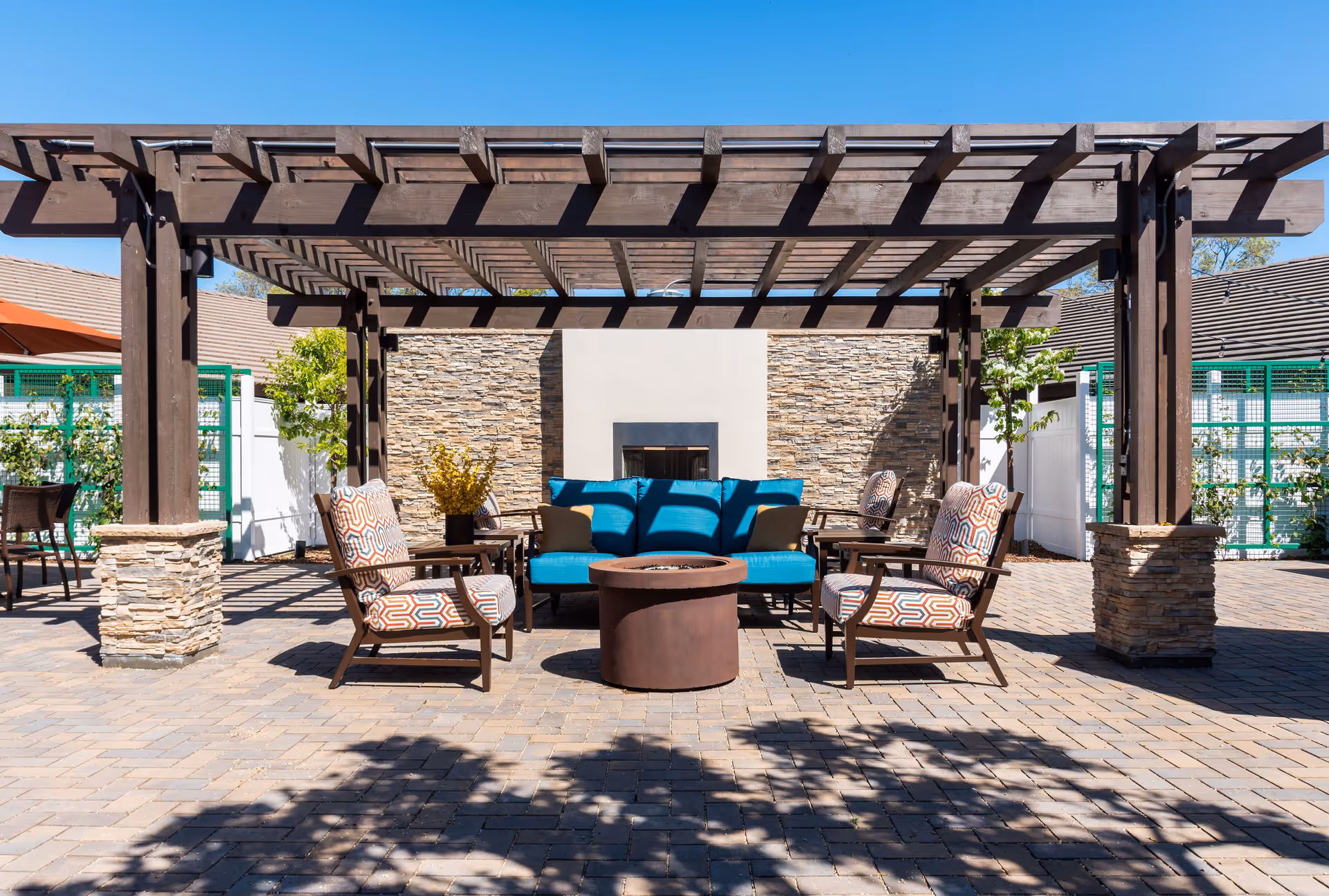 Outdoor seating area with a wooden pergola overhead, featuring a blue cushioned sofa, patterned armchairs, a round fire pit table, and a stone wall with a built-in fireplace in the background.