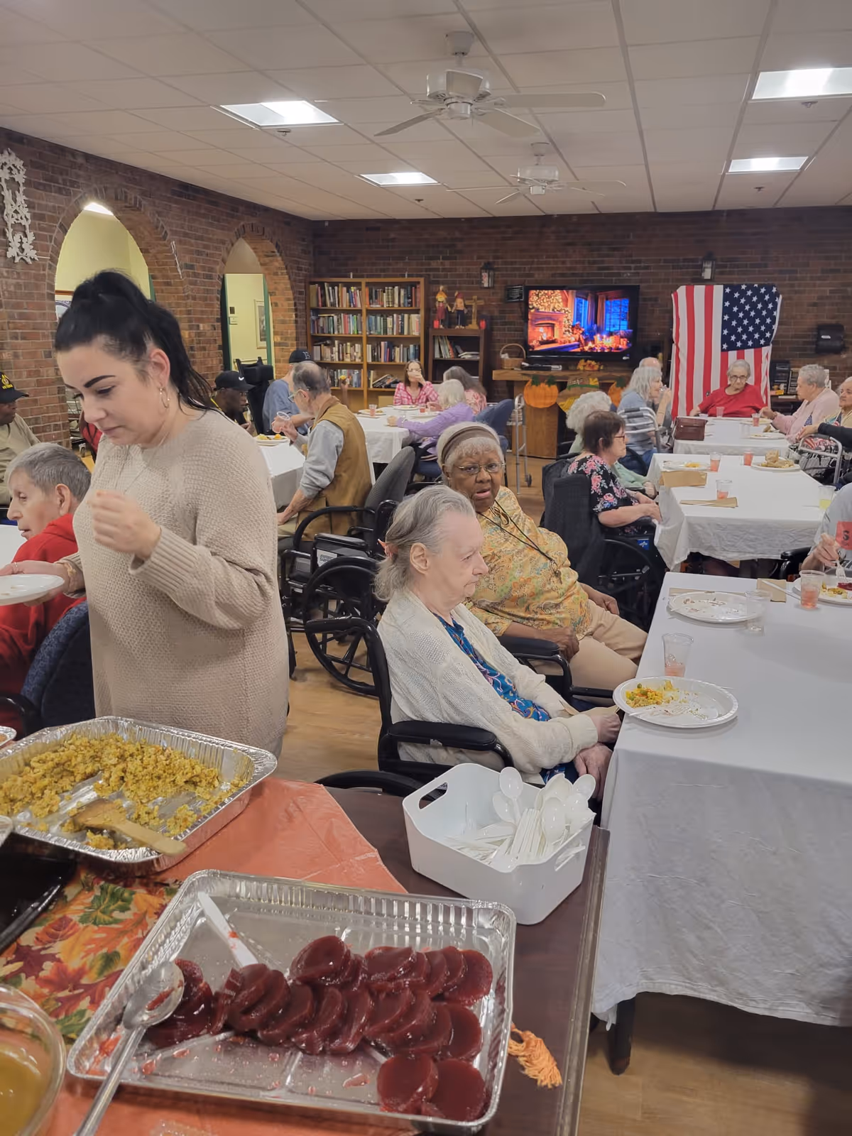 A group of elderly people seated at tables covered with white tablecloths in a communal dining area. A woman in a beige sweater is serving food from trays containing stuffing and cranberry sauce. The room has brick walls, bookshelves, and an American flag displayed near a television showing a fireplace scene. Several people are in wheelchairs, and the atmosphere appears to be a social meal gathering.