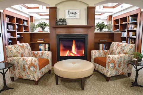 A cozy living room area with two patterned armchairs facing each other across a round beige ottoman. Behind the ottoman is a fireplace with a fire burning, framed by wooden shelves filled with books and decorative plants. Above the fireplace is a sign that reads 'home sweet home'.