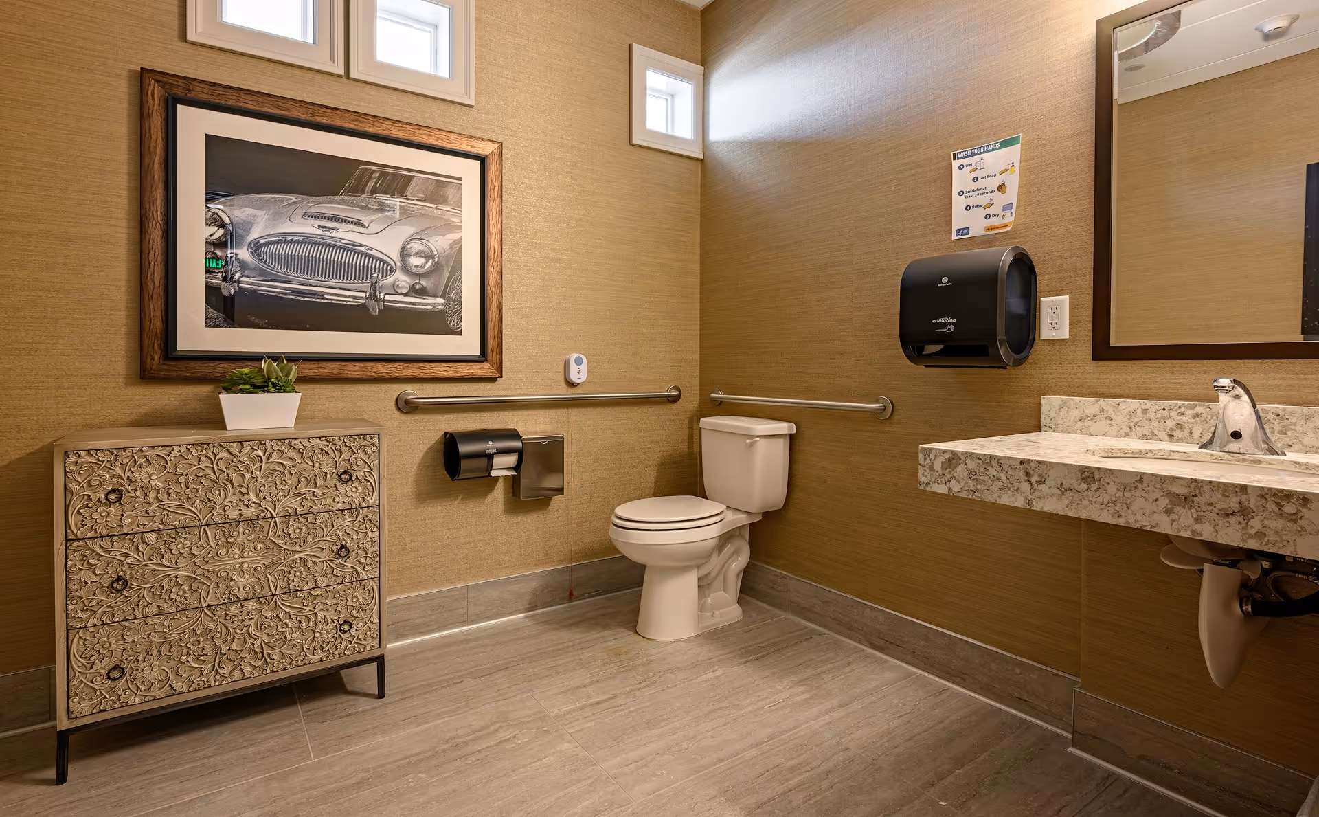 A clean and accessible bathroom with beige walls and light gray tiled floor. The room features a white toilet with grab bars on both sides, a decorative cabinet with floral carvings, a framed black and white picture of a classic car above the cabinet, a sink with a marble countertop and a large mirror, a black paper towel dispenser, and a handwashing instruction sign on the wall.