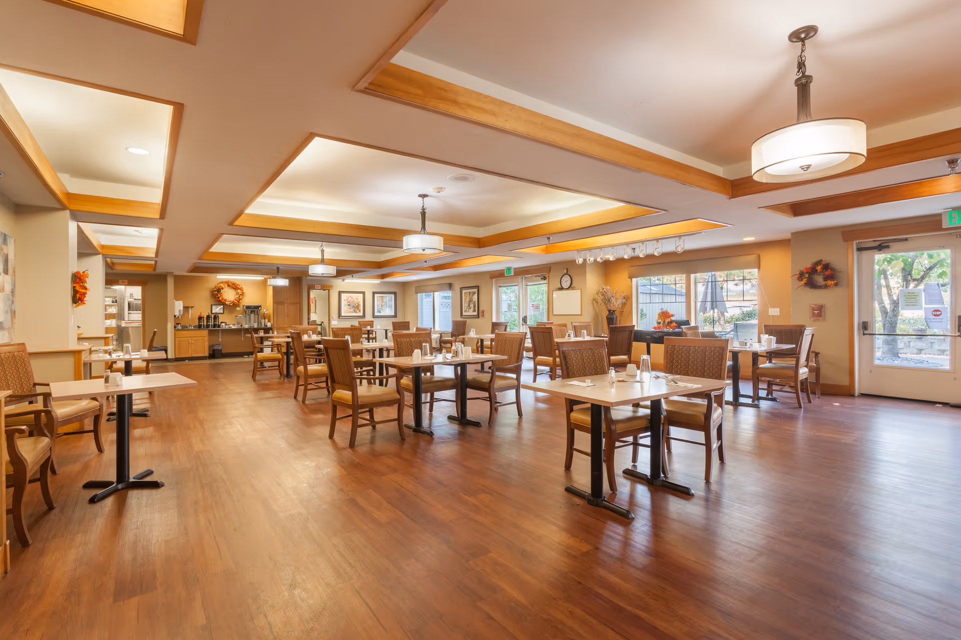 A spacious dining room in an assisted living facility with wooden floors, multiple tables and chairs arranged neatly, soft ceiling lighting, and large windows letting in natural light. The room is decorated with autumn-themed wreaths and artwork on the walls.