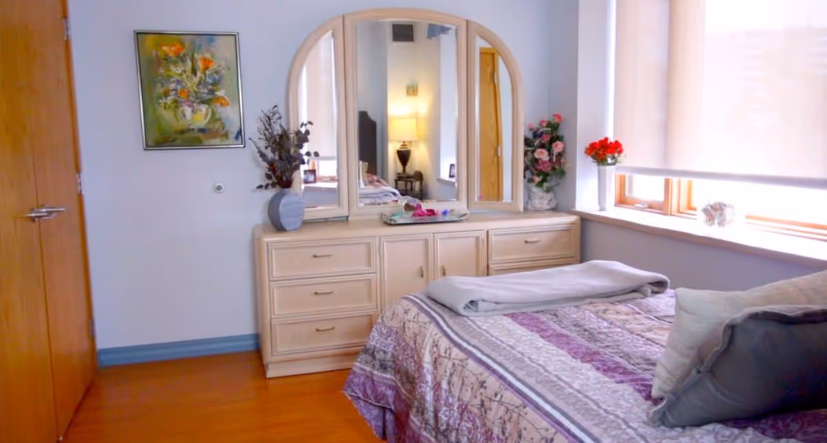 A bright bedroom with a bed covered by a patterned quilt, a dresser with a three-panel mirror and decorative vases, and a window with blinds.