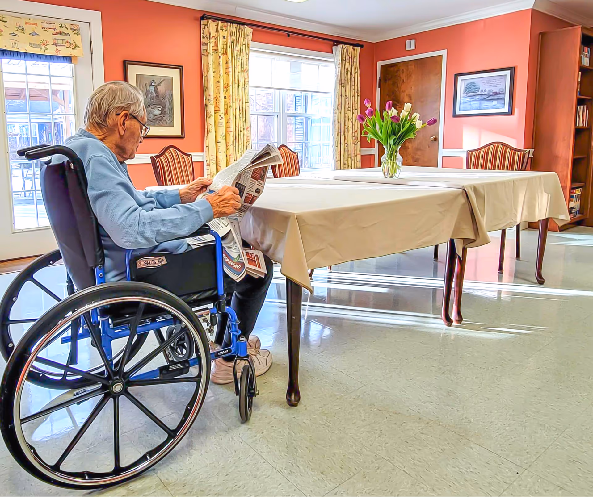 An elderly man in a wheelchair reading a newspaper at a table covered with a beige tablecloth in a brightly lit room with coral-colored walls, windows with patterned curtains, framed artwork, and a vase of tulips on the table.