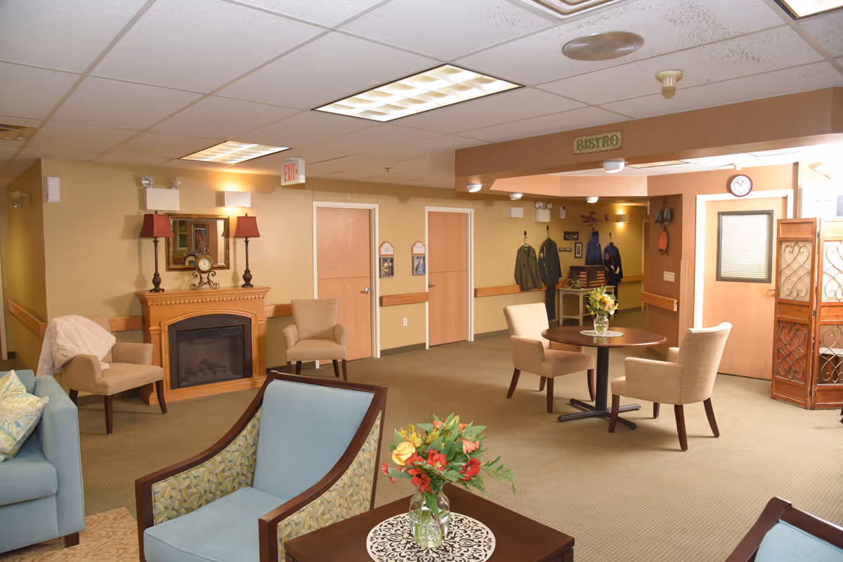 Bright furnished senior living common area with chairs, a round table, fireplace, and decorative accents.