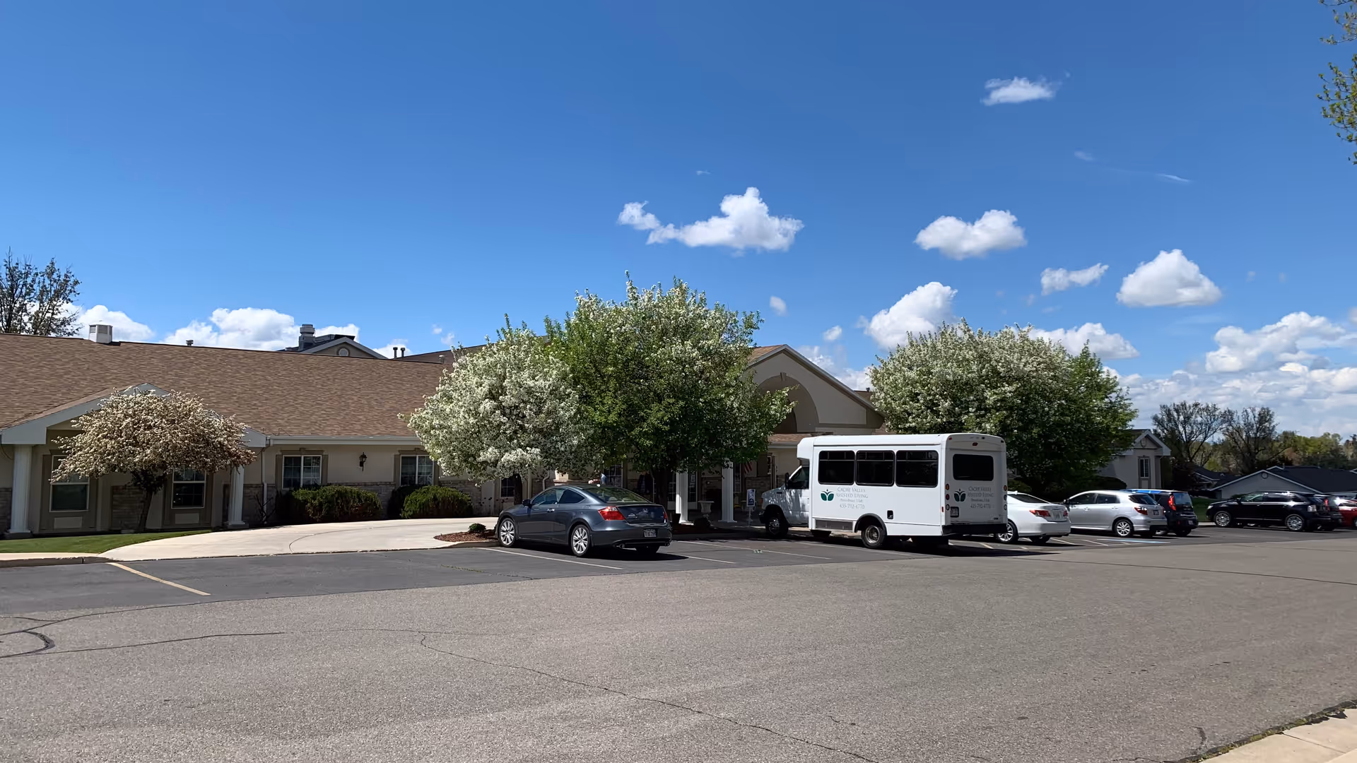 Exterior view of Cache Valley Assisted Living and Memory Care facility showing a single-story building with a brown roof, several trees with white blossoms, a parking lot with cars and a white shuttle bus, under a blue sky with scattered clouds.
