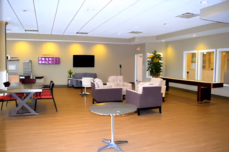 Spacious common area with wooden flooring featuring several seating arrangements including armchairs and a sofa, a round table in the foreground, a shuffleboard table on the right, and a wall-mounted TV in the background. The room is softly lit with recessed ceiling lights and has neutral-colored walls with some plants and decor.