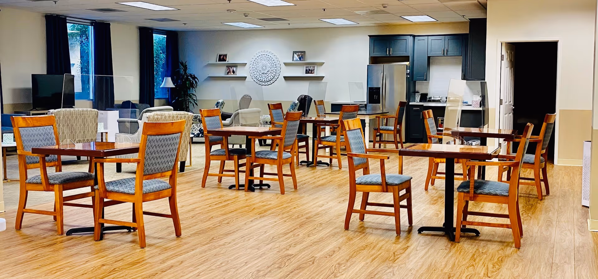 A spacious dining room with several wooden tables and chairs arranged neatly. Each table has clear protective partitions. In the background, there is a seating area with armchairs and a television. The room has large windows with dark curtains, light wood flooring, and a kitchenette with dark cabinets and a stainless steel refrigerator.