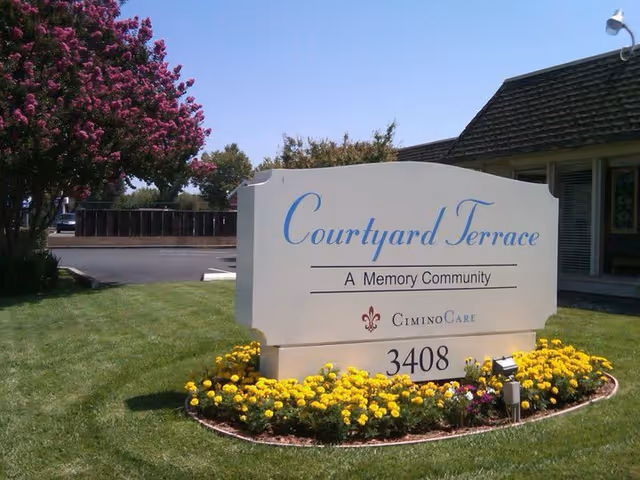 Outdoor view of a sign for Courtyard Terrace, a memory community by CiminoCare, surrounded by yellow flowers and green grass with a tree and building in the background.