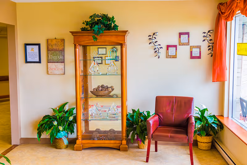 A bright seating area with a wooden glass display cabinet, a red armchair, potted plants, and wall decorations beside a window.