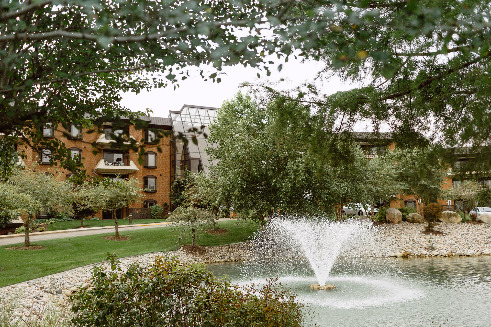 A landscaped outdoor area at Holland Home Raybrook Campus featuring a pond with a water fountain in the center, surrounded by trees and bushes. In the background, there is a multi-story brick building with balconies and large glass windows.