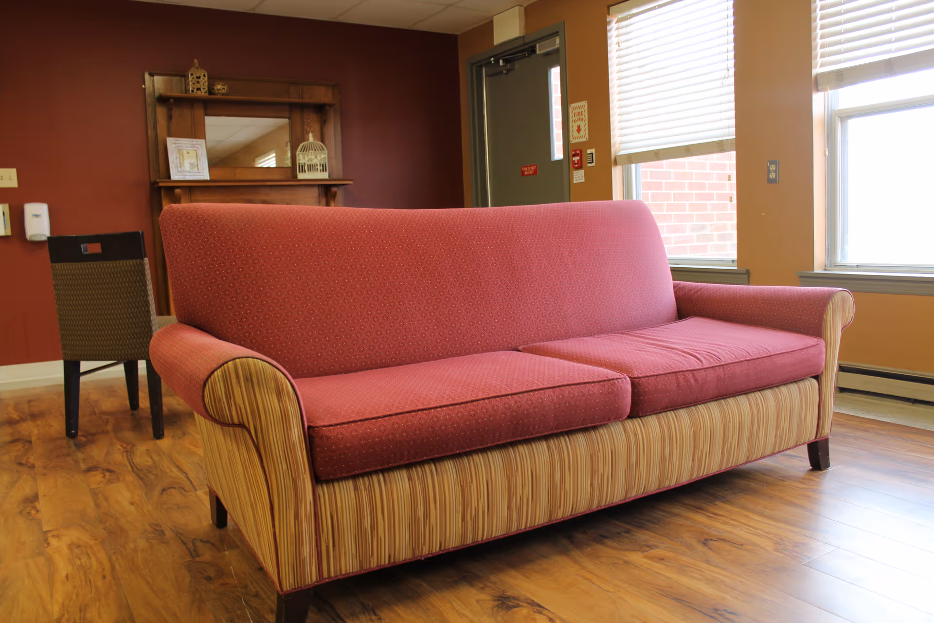 A red and beige patterned sofa is placed on a wooden floor in a room with a red accent wall and a window with blinds. Behind the sofa, there is a wooden shelf with decorative items and a chair. A door and a window letting in natural light are visible on the right side.