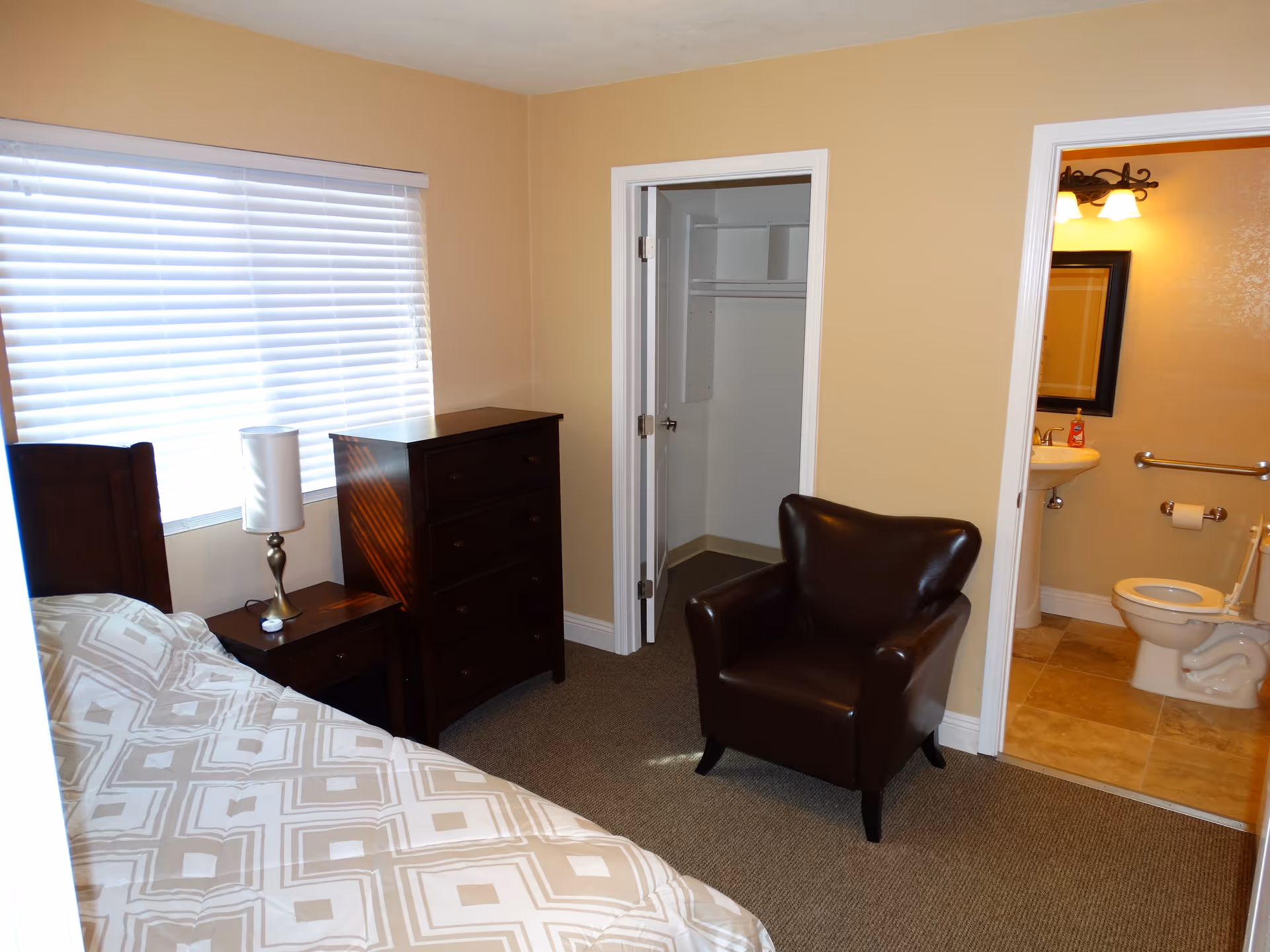 A senior care facility bedroom with a bed covered in a patterned beige and white comforter, a wooden nightstand with a lamp, a wooden chest of drawers, a brown leather armchair, an open closet, and an adjacent bathroom with a toilet, sink, mirror, and grab bars.