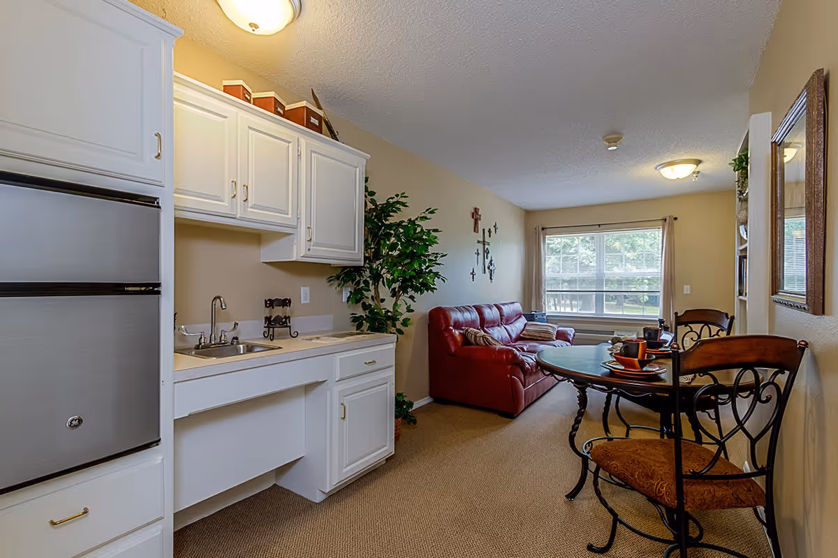 Interior view of a senior living facility room featuring a small kitchenette with white cabinets and a sink on the left, a red leather sofa against the far wall, a round dining table with two chairs on the right, and a large window with beige curtains letting in natural light.