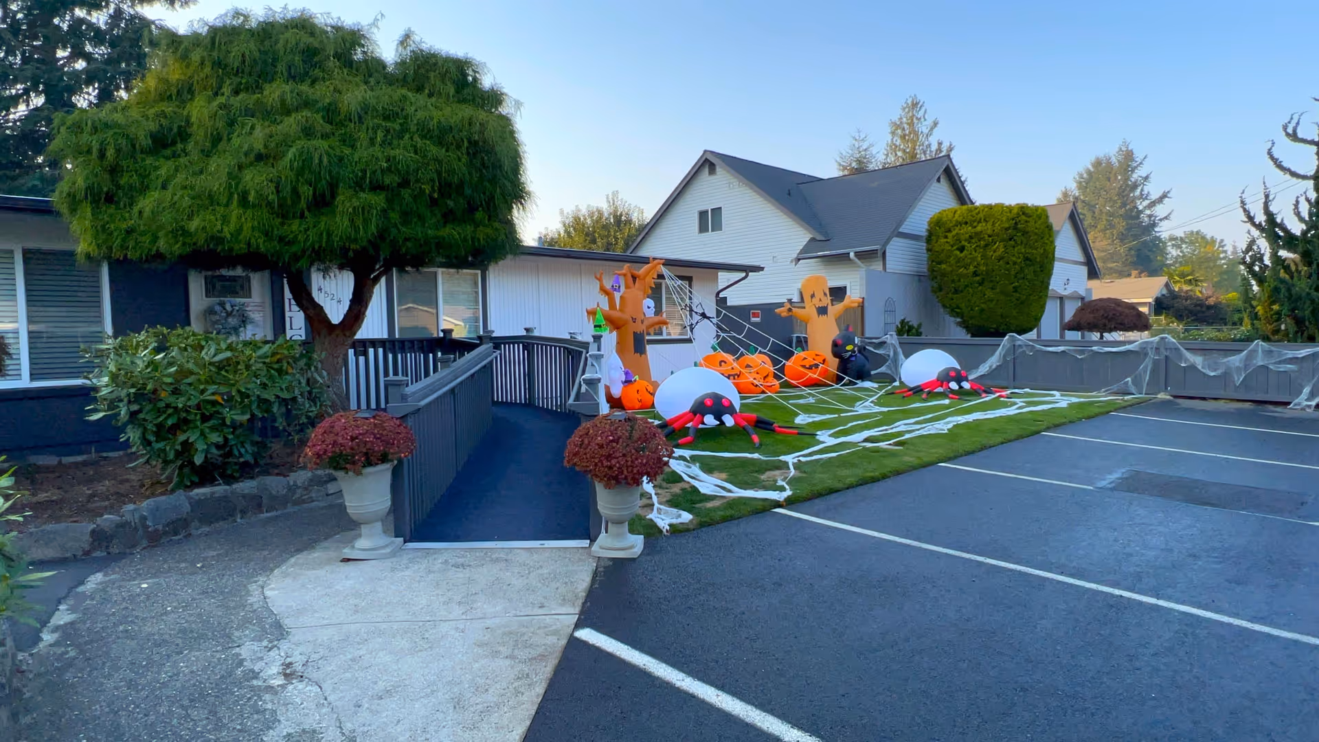 Exterior view of a senior care home with a wheelchair ramp leading to the entrance. The lawn next to the ramp is decorated with Halloween-themed inflatables including pumpkins, ghosts, spiders, and spider webs. There are houses and trees in the background under a clear sky.