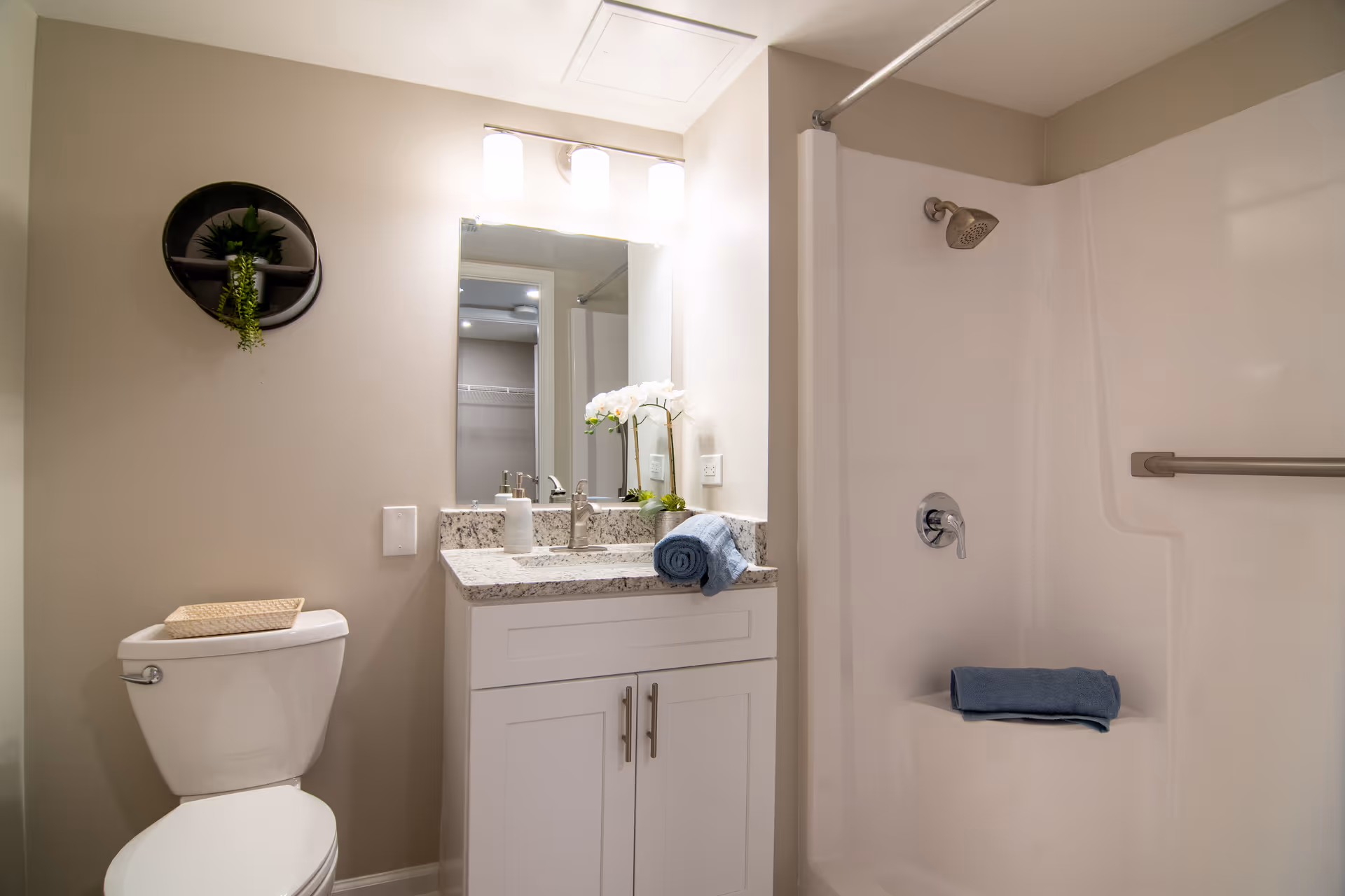 A clean and modern bathroom featuring a white toilet with a wicker basket on top, a white vanity with granite countertop, a sink with a soap dispenser, a rolled blue towel, and a small plant. Above the vanity is a mirror with three lights. To the right is a white shower area with a built-in seat, a showerhead, a grab bar, and another rolled blue towel on the seat. A round wall shelf with a small plant is mounted on the left wall.