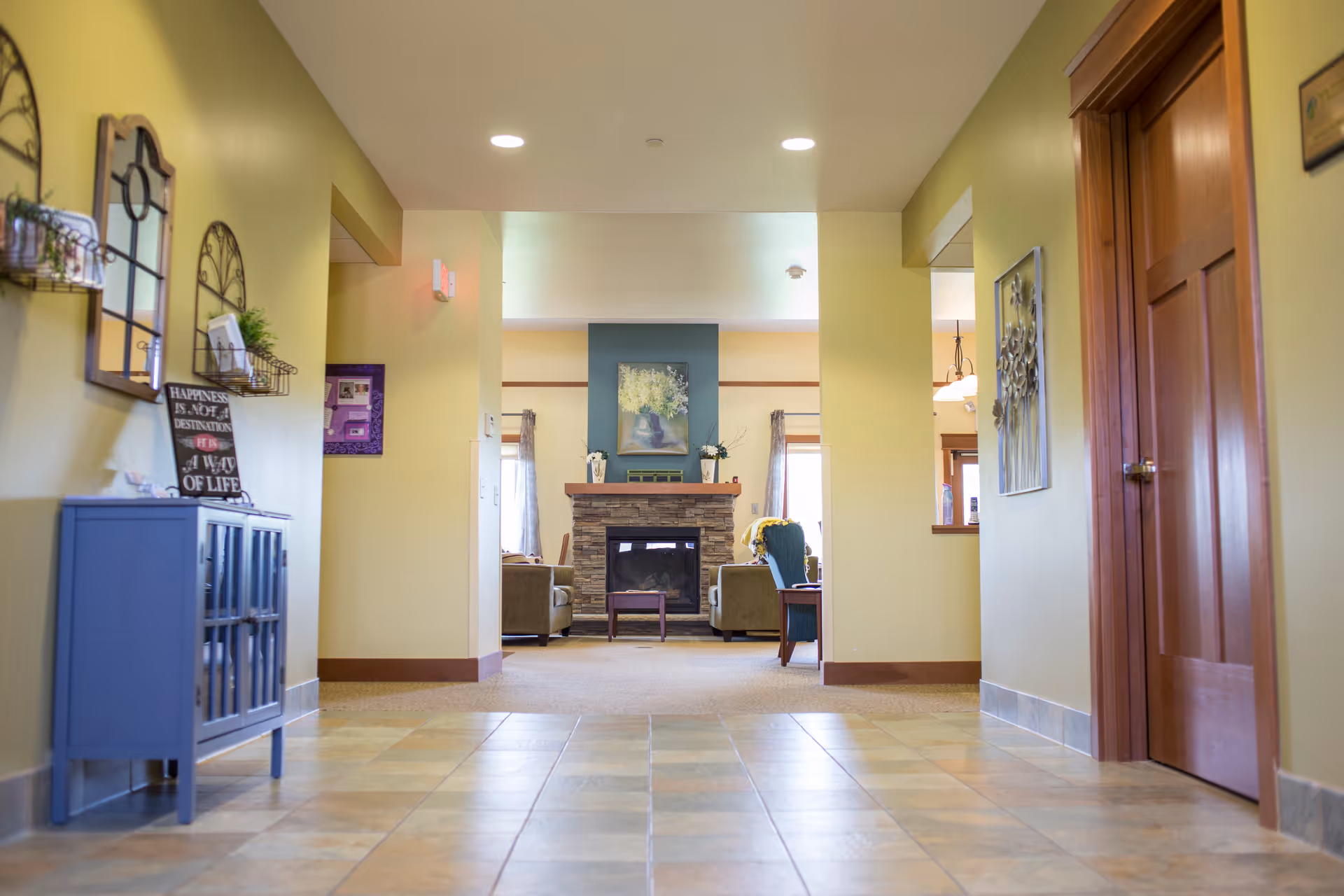 View down a tiled hallway leading to a cozy living room area with a stone fireplace, armchairs, and a painting above the mantel. The hallway walls are painted light green with decorative wall hangings and a small blue cabinet on the left side. There is a wooden door on the right and a window opening to another room on the right side.