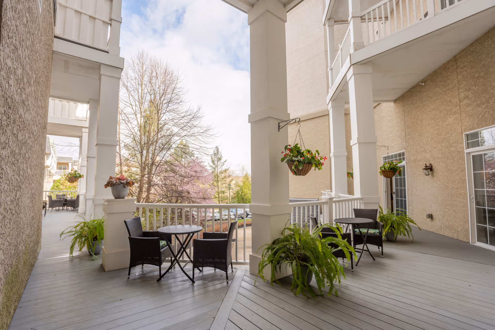 Outdoor covered patio area with round tables and black wicker chairs, decorated with hanging flower pots and potted plants. The patio overlooks trees and a parking area under a partly cloudy sky.