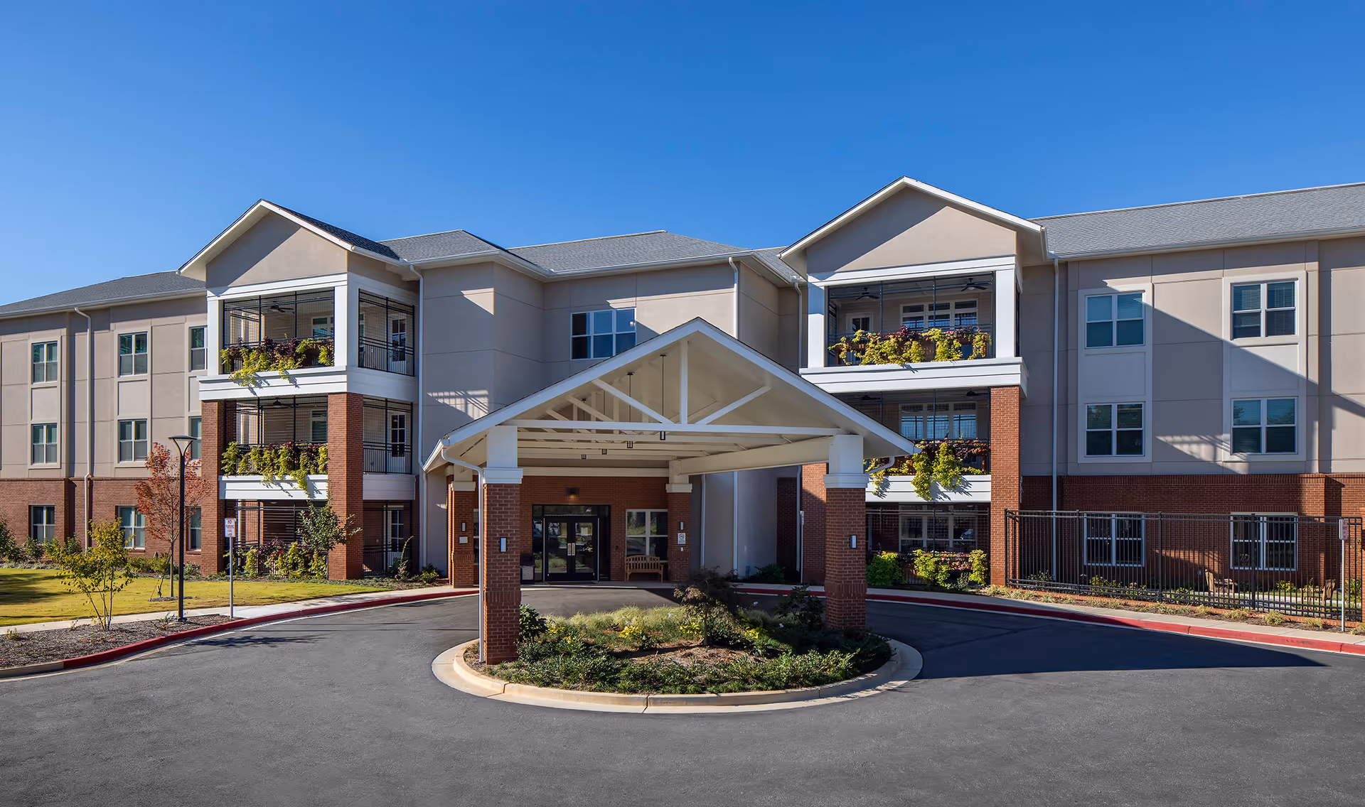 Front exterior view of a multi-story senior living facility building with a covered entrance, balconies with plants, and a circular driveway under a clear blue sky.