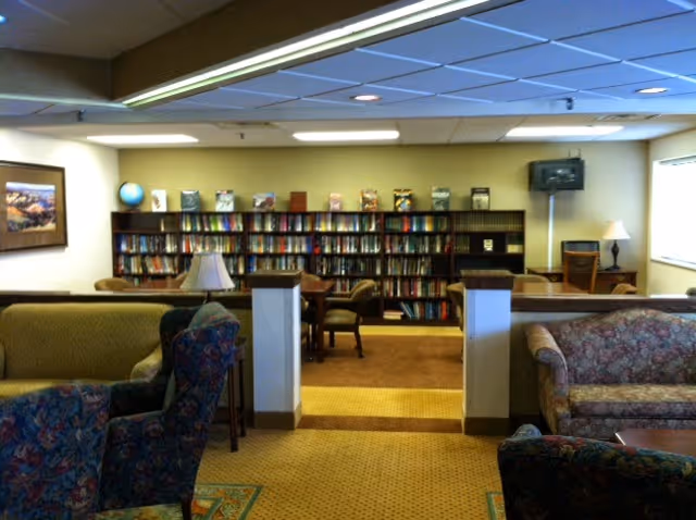A cozy senior living common area with patterned armchairs and sofas arranged around small tables. In the background, there is a large bookshelf filled with books and a small TV mounted on the wall. The room is well-lit with ceiling lights and has a warm, inviting atmosphere.