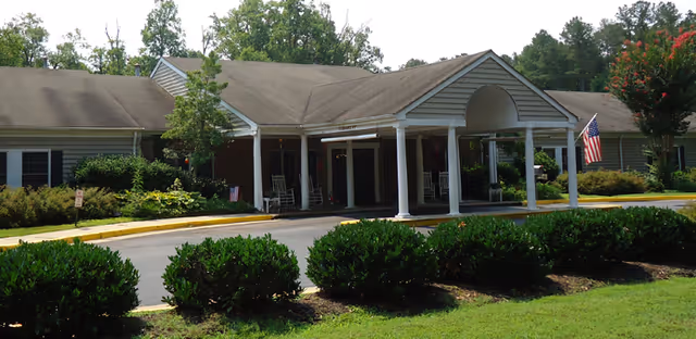 Exterior view of a single-story assisted living center building with a covered entrance supported by white columns. The building has a gray roof and beige siding with black shutters on the windows. There are bushes and trees surrounding the building, and an American flag is displayed near the entrance.
