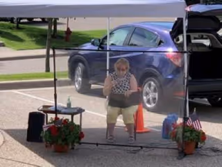 An elderly woman sitting on a chair behind a clear protective barrier outdoors in a parking lot. She is under a white canopy tent, with potted flowers on either side of the barrier. A blue SUV with its trunk open is parked behind her, and there is a traffic cone nearby.