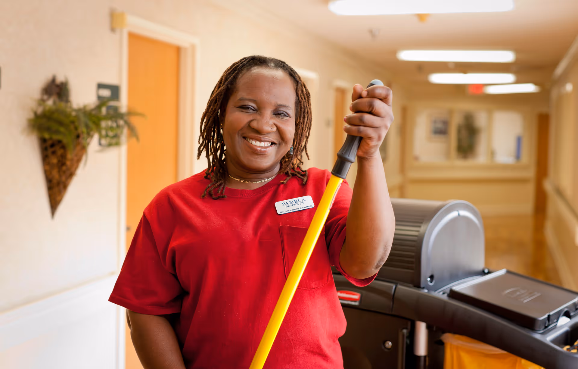 A smiling woman wearing a red shirt and a name tag that reads 'Pamela' stands in a well-lit hallway holding a yellow mop handle. Behind her is a janitorial cart with cleaning supplies. The hallway has beige walls, doors, and some wall decorations.