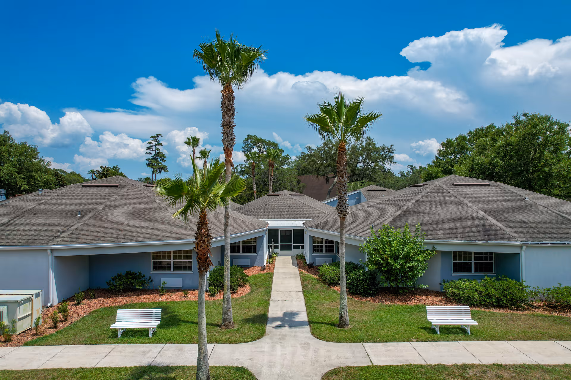 Exterior view of a single-story building with a gray roof and light blue walls, surrounded by green grass, palm trees, and bushes under a partly cloudy blue sky. Two white benches are placed on either side of a concrete walkway leading to the building entrance.