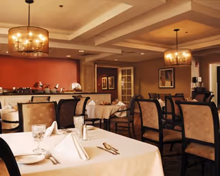 A warmly lit dining room with multiple tables covered in white tablecloths, each set with folded napkins, glassware, and silverware. The room features upholstered chairs with patterned backs, two large modern chandeliers hanging from a coffered ceiling, and framed artwork on the walls. There is a buffet area along one wall with plates and serving dishes.