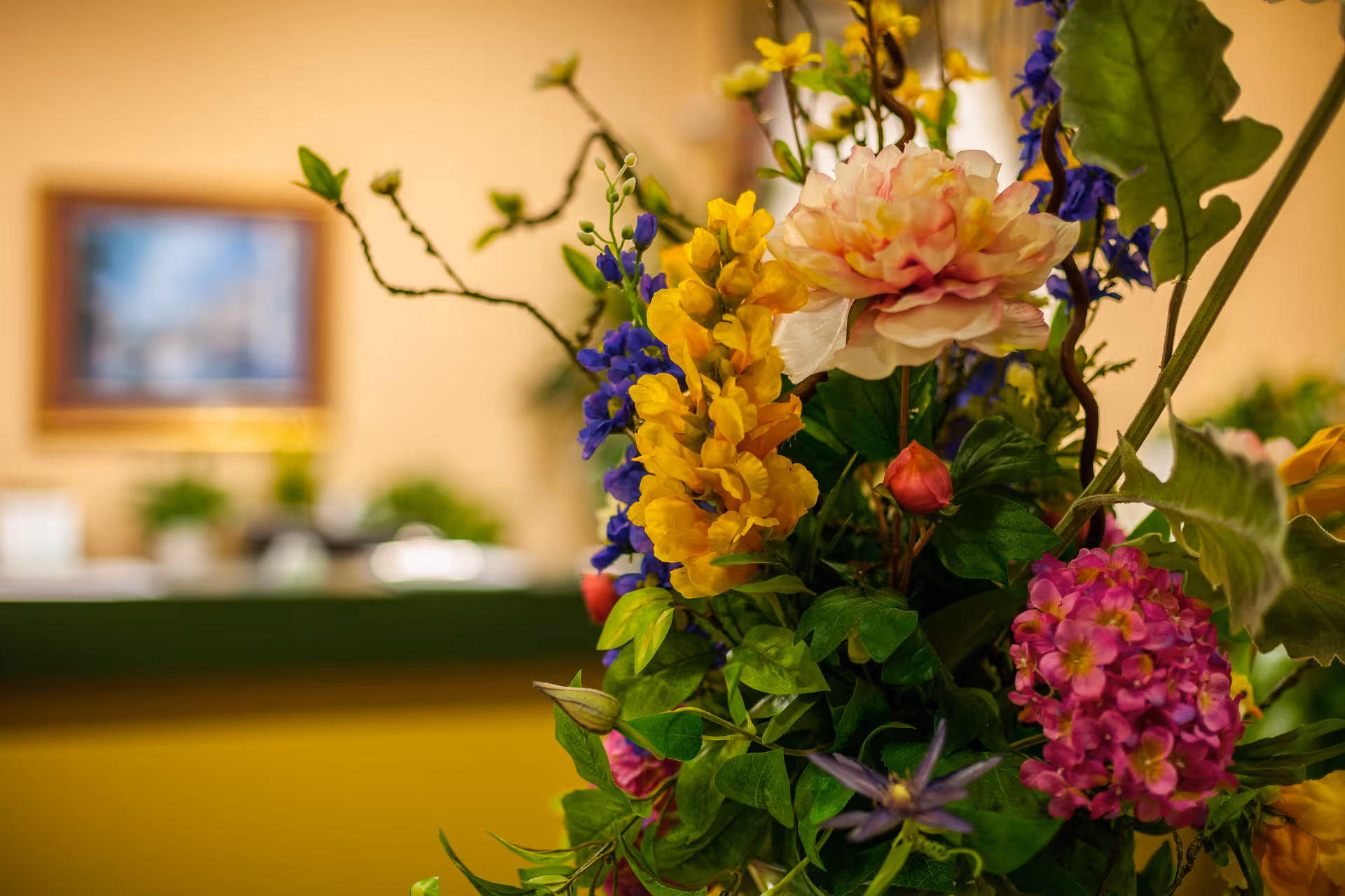 Close-up of a colorful floral arrangement in a lobby with a blurred reception desk and framed artwork in the background.
