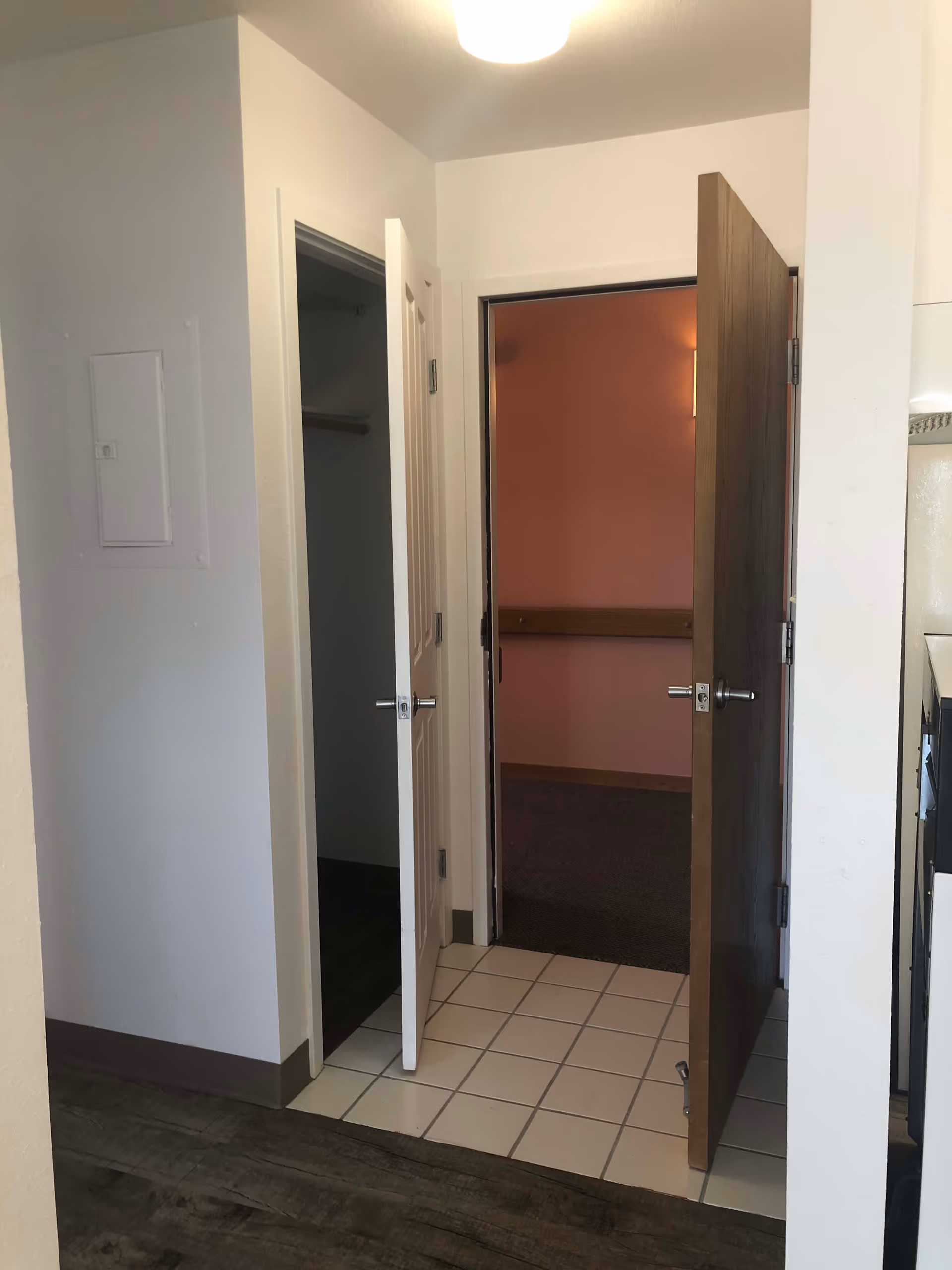 Interior view showing two open doors in a senior living facility. One door reveals a small closet with a hanging rod, and the other door leads to a room with pink walls and a wooden handrail. The floor transitions from wood to white tiles near the doors.