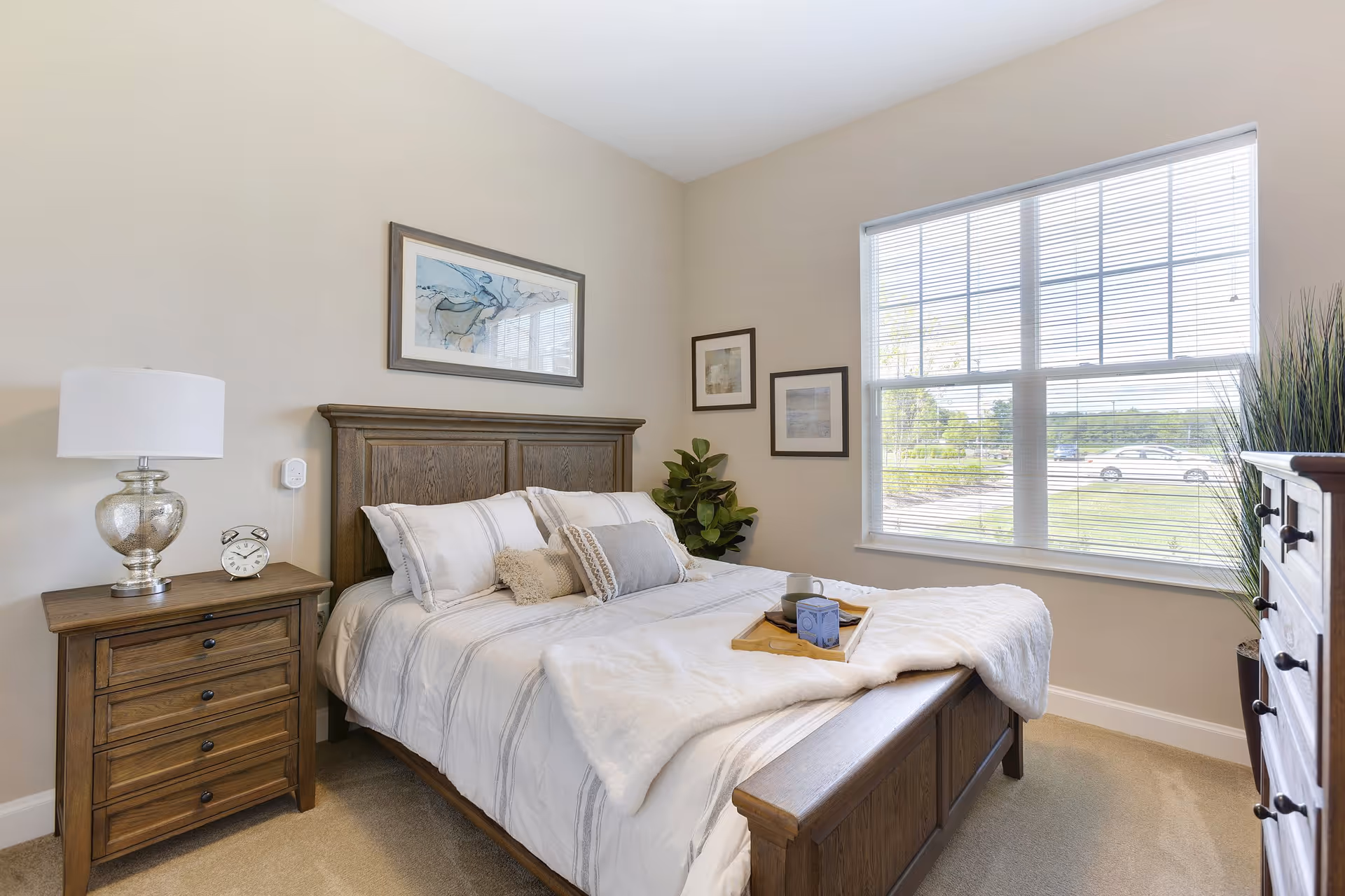 A bright bedroom with a large window letting in natural light. The room features a wooden bed with white and gray bedding, a wooden nightstand with a silver lamp and an alarm clock, framed artwork on the walls, a green plant in the corner, and a wooden dresser. A tray with a cup and a box is placed on the bed.