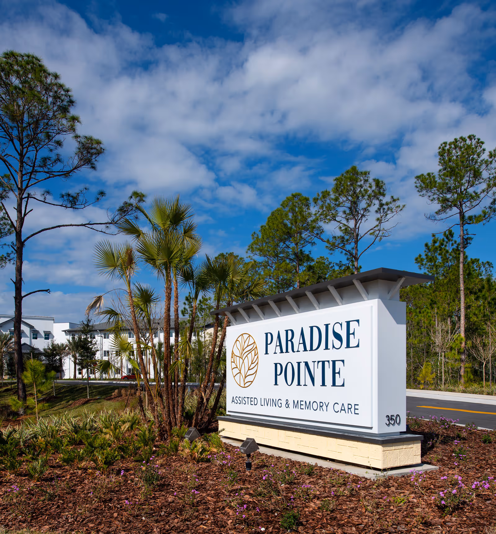 Outdoor view of the entrance sign for Paradise Pointe Assisted Living & Memory Care, surrounded by landscaped plants and trees under a partly cloudy blue sky.