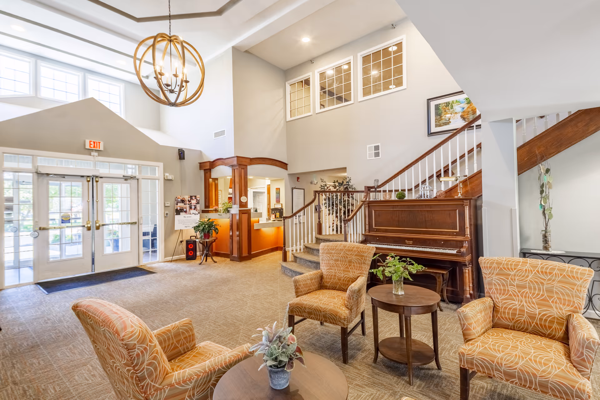 Bright senior living lobby with seating, a piano, a reception desk, and a staircase near the entrance.