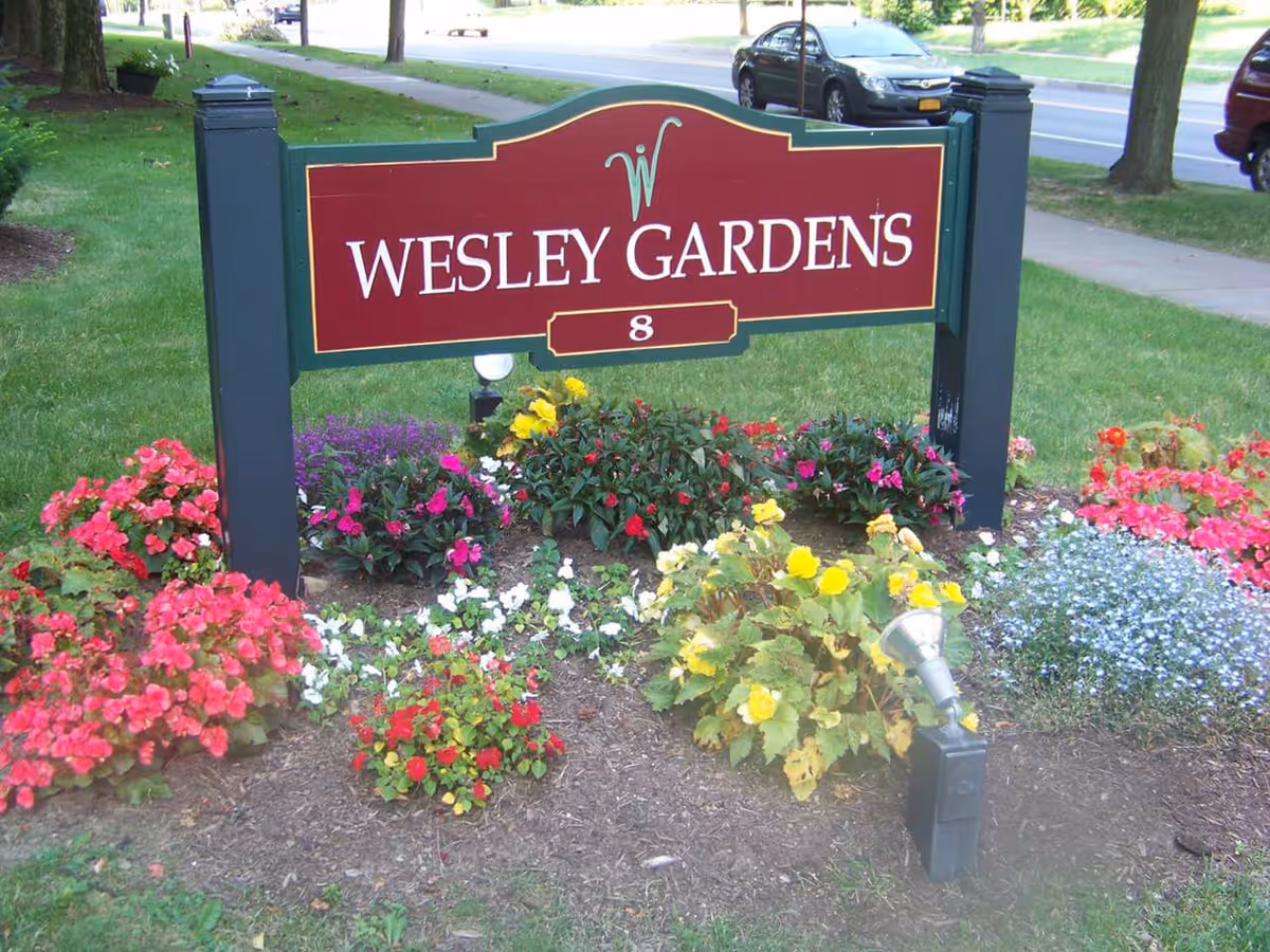 A red and green sign with the text 'Wesley Gardens' and the number 8, surrounded by a flower bed with colorful flowers including red, yellow, white, and purple blooms. The sign is located on a grassy area near a sidewalk and a street with parked cars.