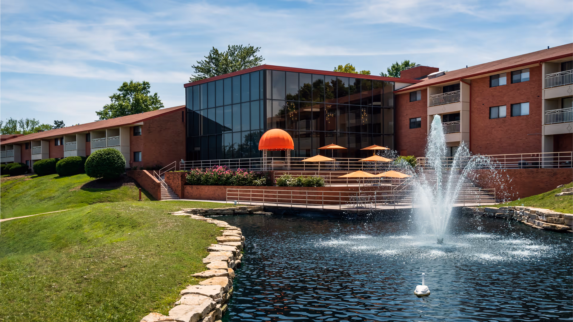 Exterior view of a senior living facility named Garden Villas featuring a large pond with a water fountain in the foreground, a grassy area with stone edging, and a building with large glass windows and orange umbrellas on a patio.