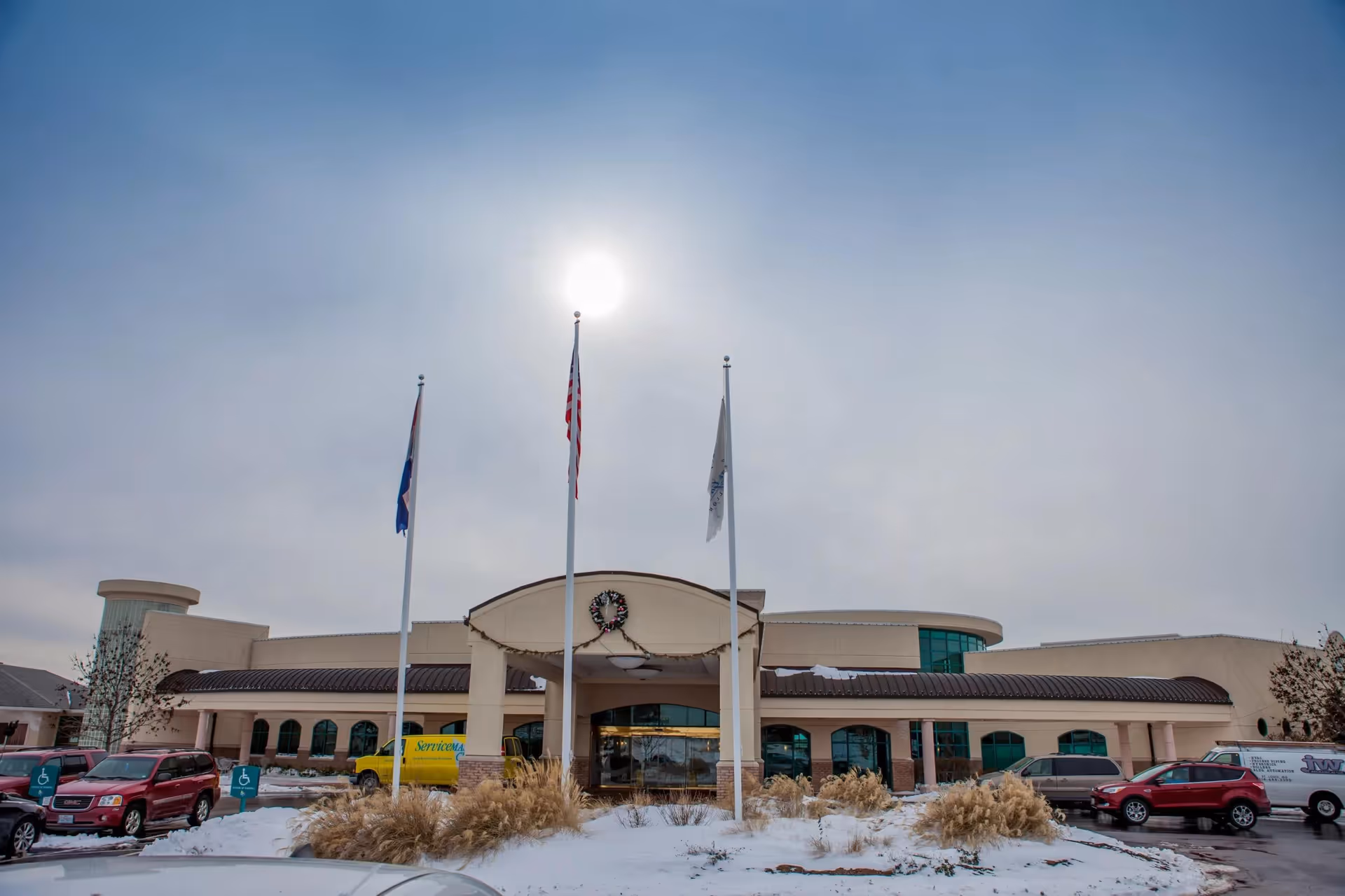 Front exterior of a senior living facility with flagpoles, an entrance canopy, parked cars and snow in the foreground under a cloudy sky.