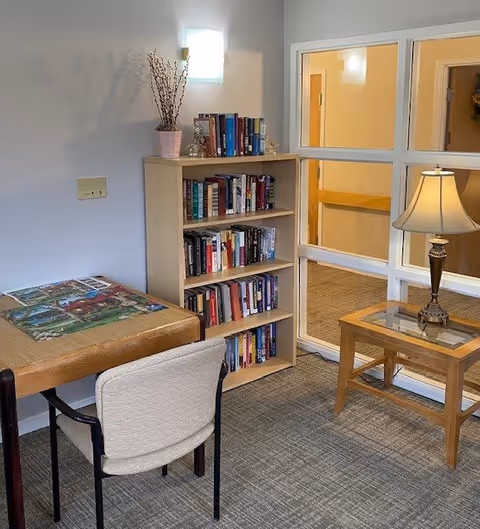 A cozy interior corner of a senior living facility featuring a wooden table with a puzzle in progress, a cushioned chair, a bookshelf filled with books, a small wooden side table with a lamp, and a windowed wall showing a hallway outside.
