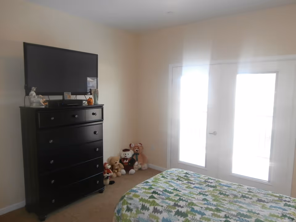 Bedroom with a patterned bed, a black dresser topped by a wall-mounted TV, stuffed animals in the corner, and bright double glass doors.