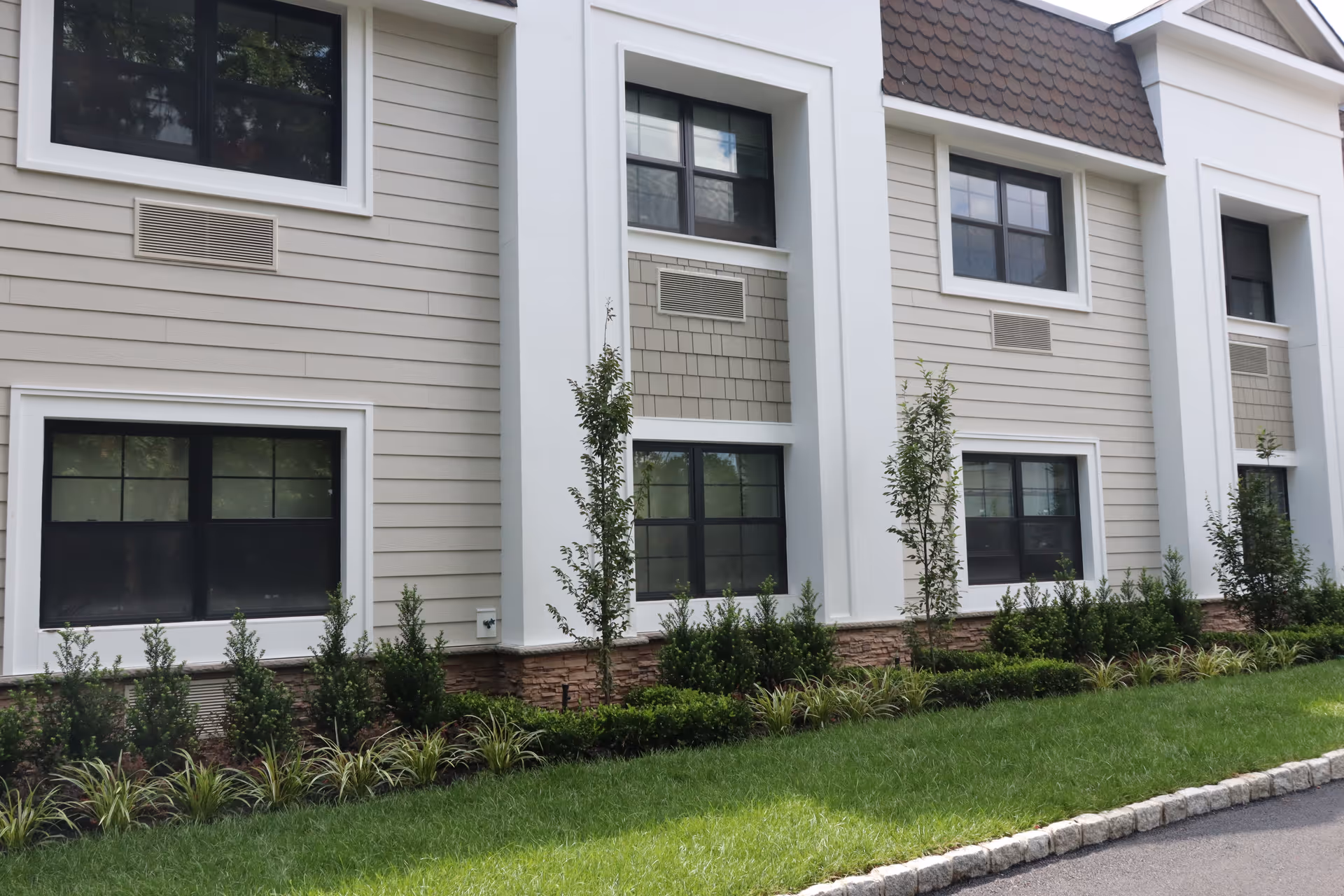 Exterior view of a building with beige siding, white trim, and multiple windows. There are small trees and shrubs planted along the base of the building with a well-maintained lawn and a paved pathway in front.