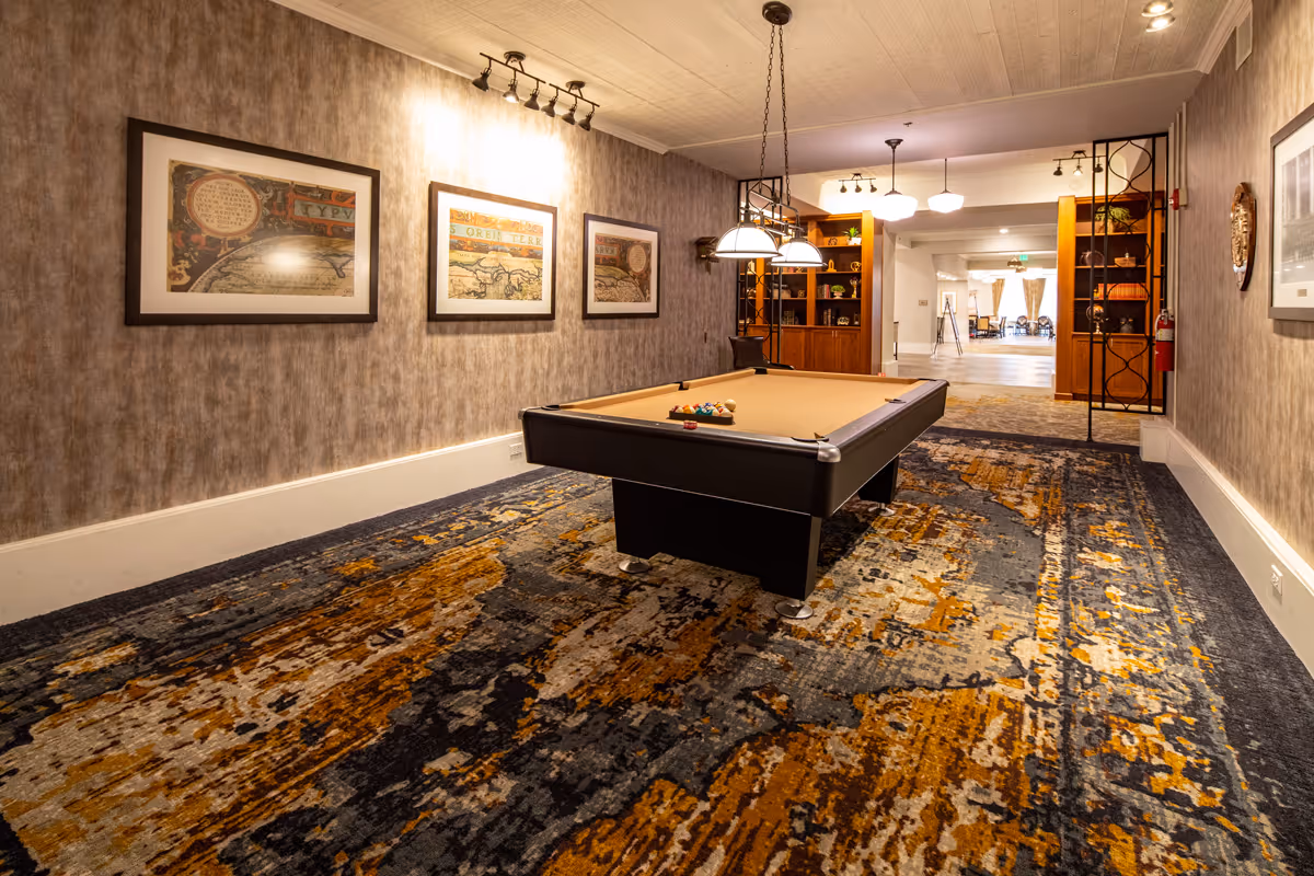 Interior view of a recreational room featuring a pool table with billiard balls arranged on it. The room has patterned carpet flooring in shades of blue, orange, and gray, textured wallpaper, and three framed artworks on the left wall. Overhead lights hang above the pool table, and wooden shelves with decorative items are visible in the background near an open doorway leading to another room with chairs and windows.