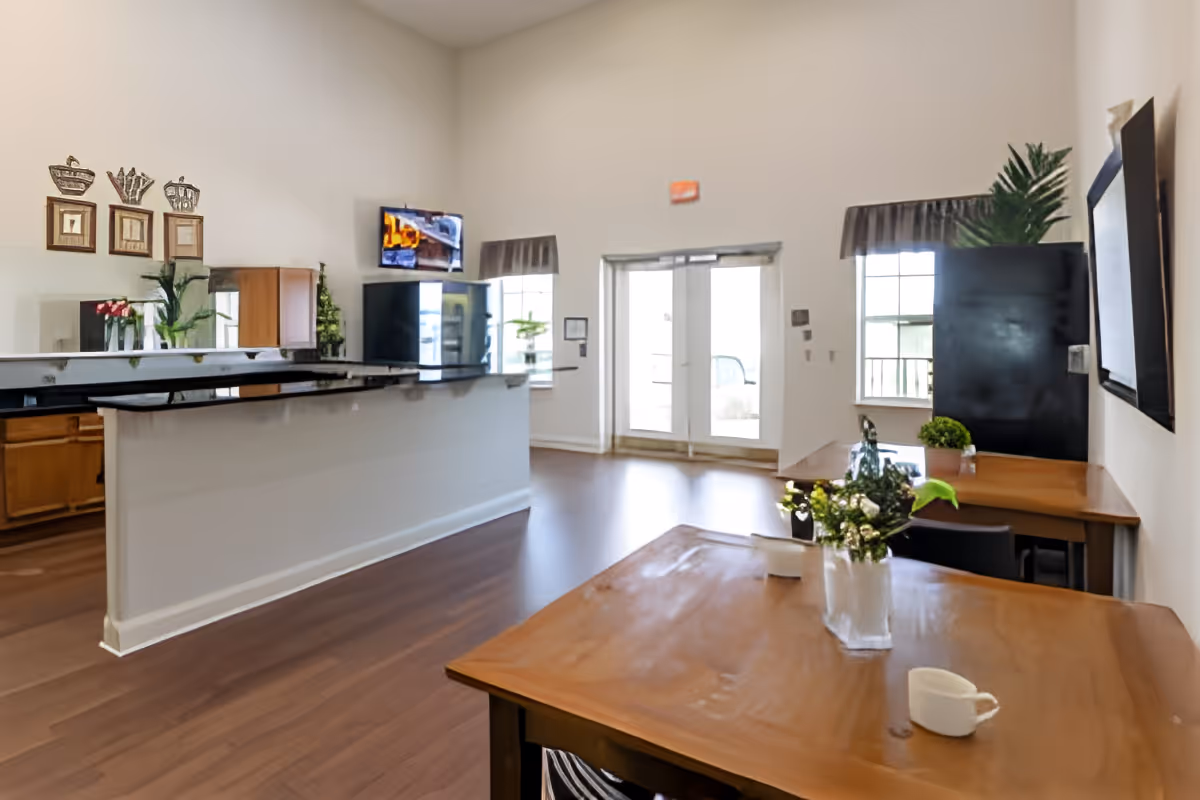 Interior view of a bright common area in a senior living facility with wooden flooring, a long counter with a black countertop, a mounted TV, two windows with curtains, a door leading outside, and wooden tables with small plants and a white cup on them.