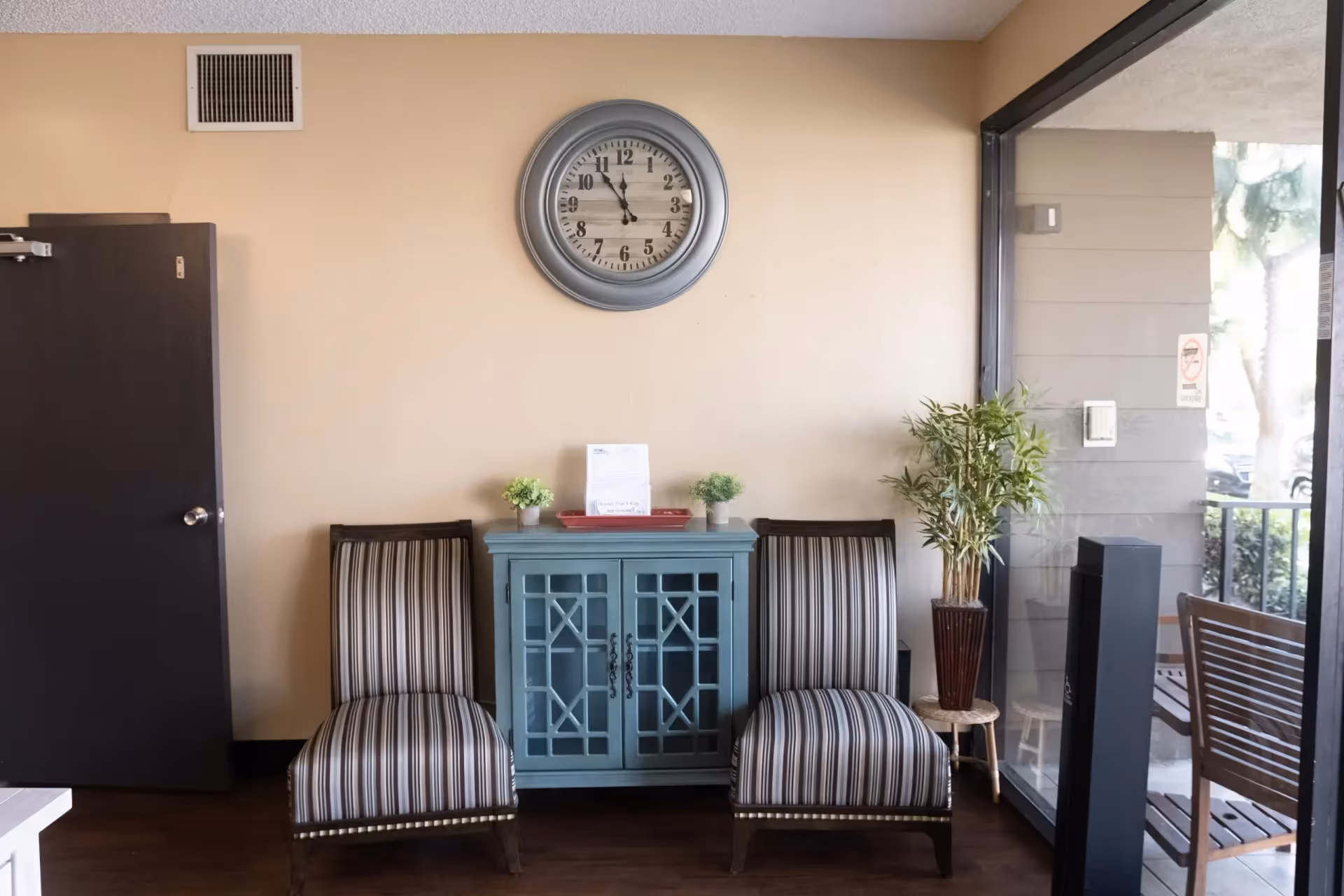 A small seating area with two striped upholstered chairs on either side of a blue cabinet with glass doors. On top of the cabinet are two small potted plants and a document holder. Above the cabinet is a round wall clock showing the time as 11:55. To the right is a large glass window with a view of an outdoor patio area with wooden chairs and a potted plant on a small stool. A closed dark door is on the left side of the image.