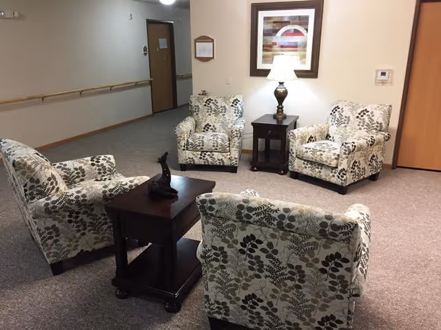 A sitting area in an assisted living facility with four floral-patterned armchairs arranged around two dark wooden side tables. One table has a decorative sculpture and the other has a lamp with a framed abstract artwork hanging above it. The room has beige carpet, light-colored walls, and a hallway with doors in the background.