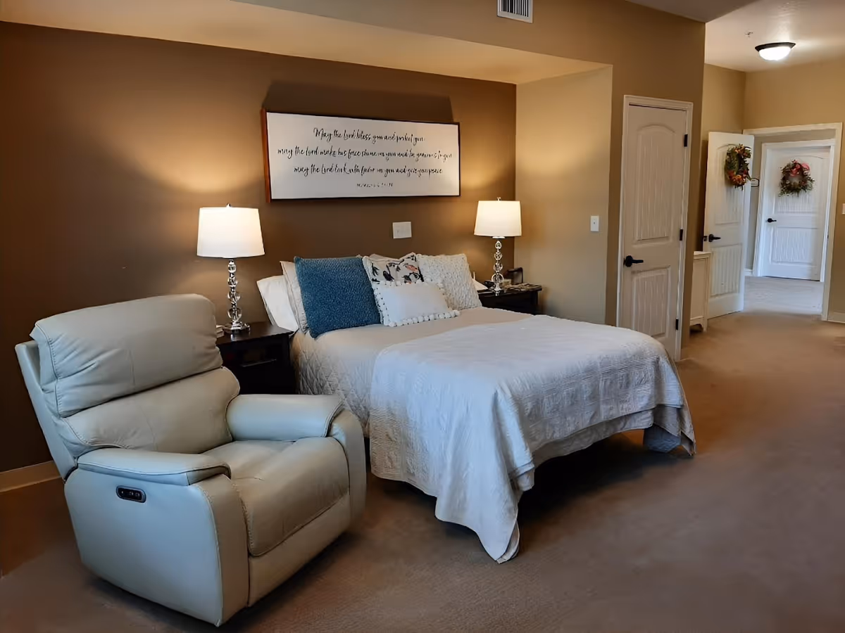 A cozy bedroom with a bed covered in a white quilt and several pillows, flanked by two nightstands each with a lamp. A comfortable beige recliner chair is positioned to the left of the bed. The walls are painted in warm tones, and there is a framed inspirational quote hanging above the bed. In the background, there are two doors, one open showing another room with wreaths hanging on the doors.