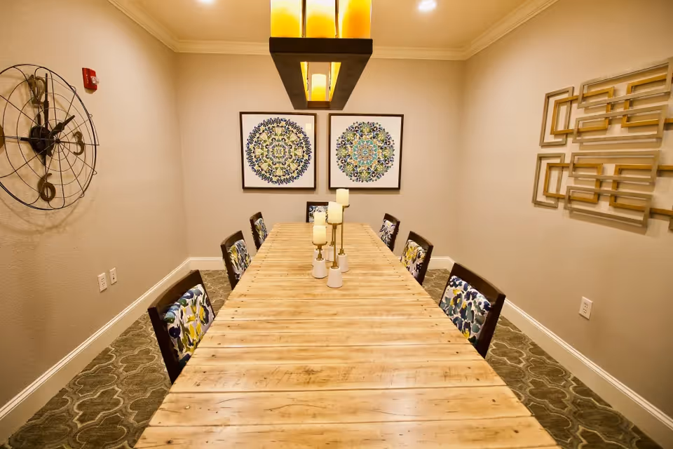 A long wooden dining table with eight chairs featuring floral patterned cushions in a well-lit room. The table is decorated with several candle holders with candles. On the walls, there is a large decorative clock on the left, two framed circular patterned artworks at the far end, and a geometric wall sculpture on the right.