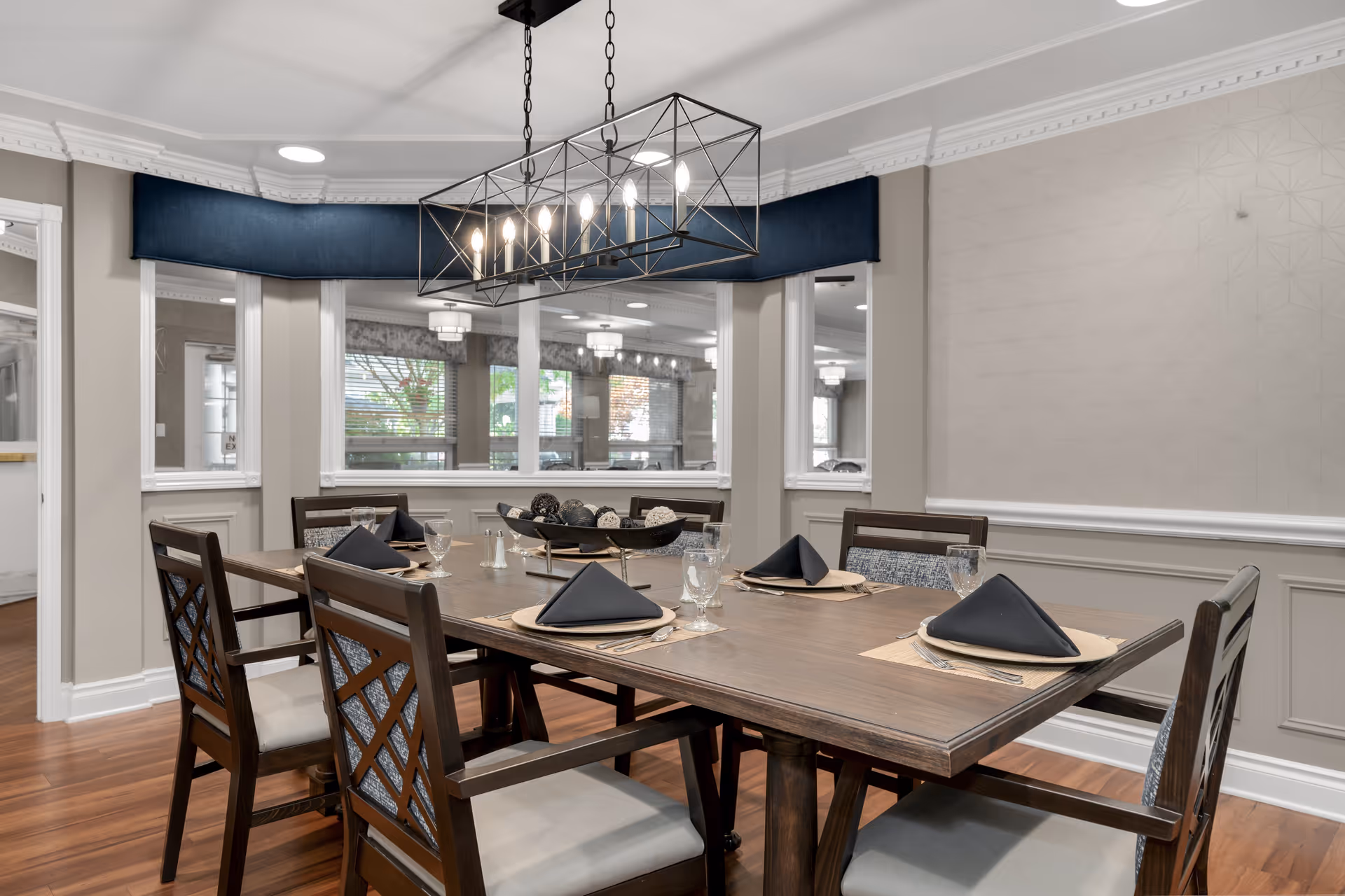 A dining room with a wooden table set for six people, each place setting includes a black folded napkin, a glass, and silverware. The room features wooden chairs with cushioned seats, a modern rectangular chandelier with exposed bulbs hanging above the table, and large windows with a view into another room. The walls are painted in neutral tones with decorative molding and a patterned wallpaper accent.